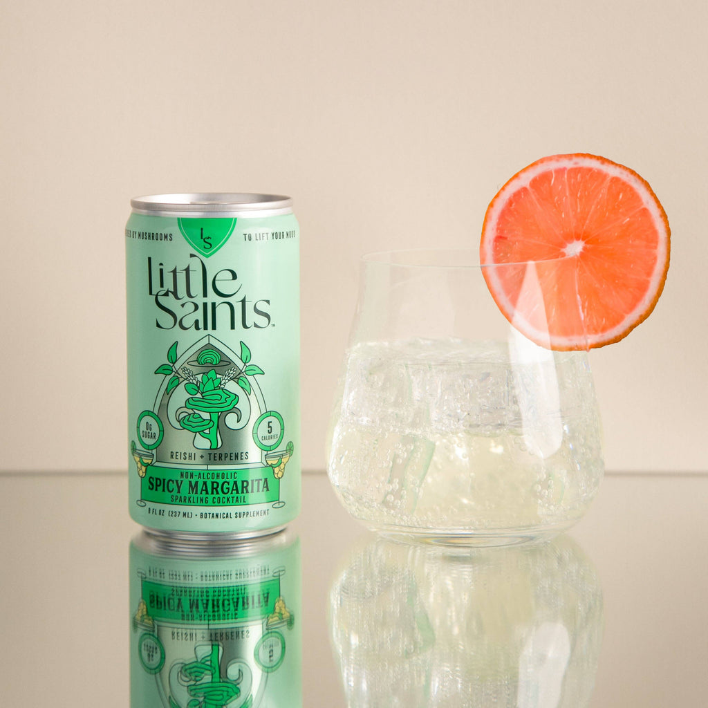 A can of Little Saints Spicy Margarita Adaptogenic Mocktail sits beside a glass of ice and clear liquid, garnished with a grapefruit slice on the rim, all set against a plain beige background.