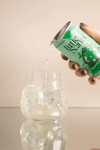 A hand pours a clear drink from a mint-green Little Saints Spicy Margarita Adaptogenic Mocktail can into an ice-filled glass; this non-alcoholic beverage, crafted with reishi mushroom, sits on a reflective surface against a neutral background.