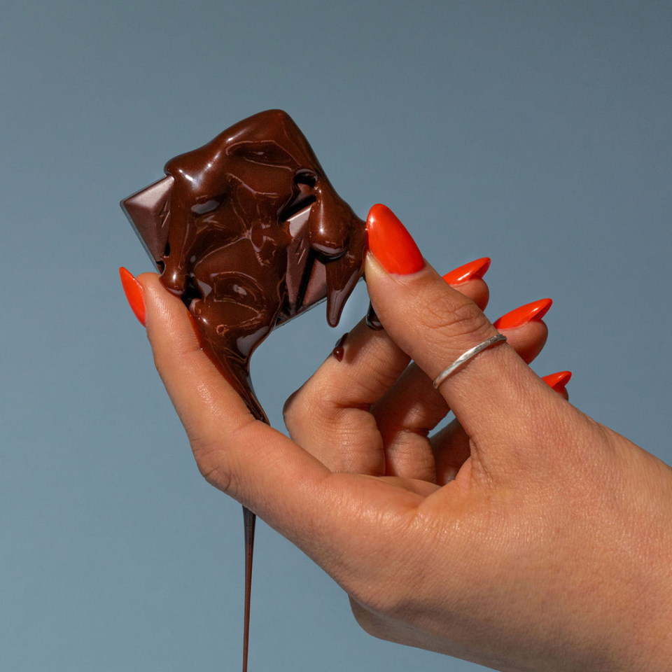 A hand with long, bright red nails holds a melting Fun Guy Chill Chocolate bar—known as a natural mood enhancer—with chocolate dripping down the fingers, set against a plain blue-gray background.