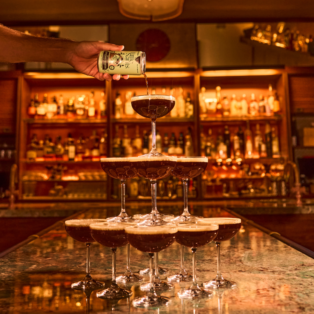 A hand pours a can of Little Saints Espresso Martini Adaptogenic Mocktail into the top coupe glass of a champagne tower filled with dark liquid, set on a bar with bottles in the background.