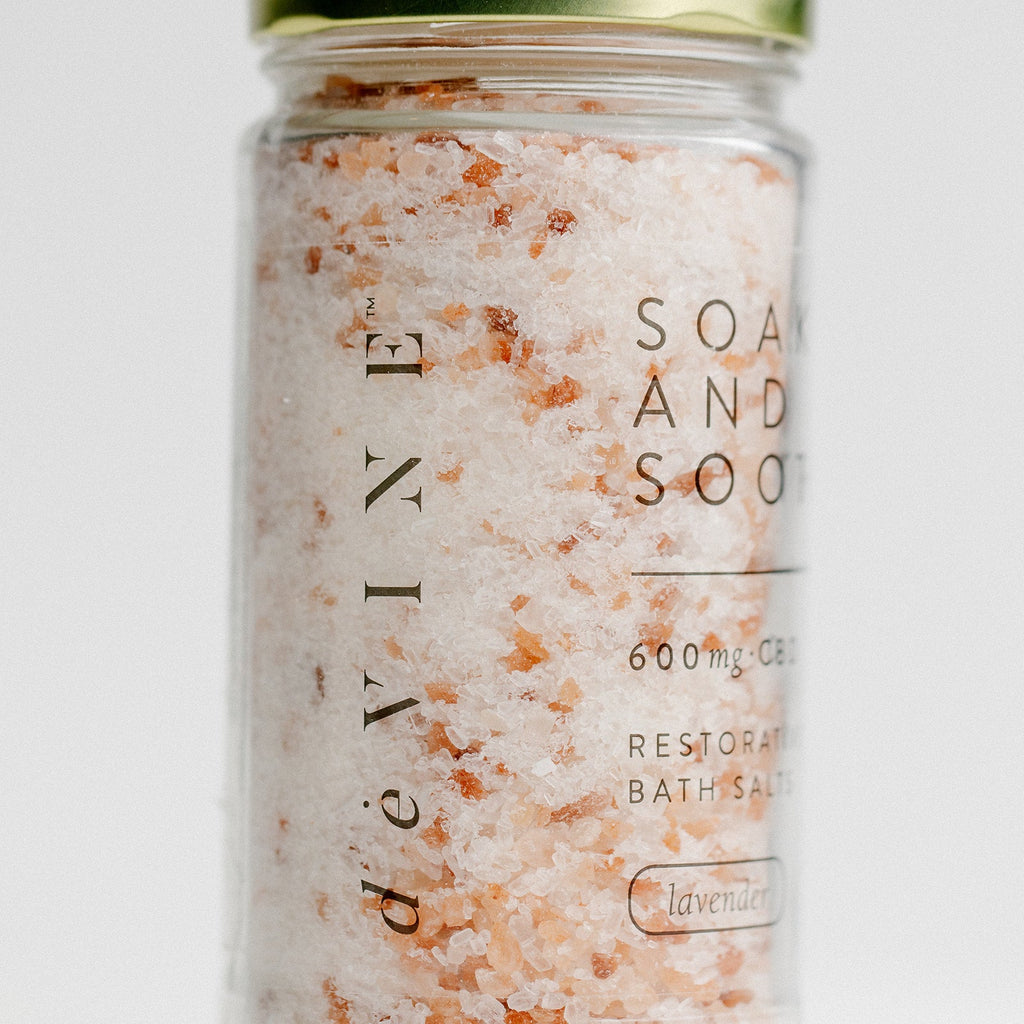 A clear jar of DeVINE Wellness Soak & Soothe Bath Salts holds pink and white bath soak with 600mg CBD, lavender essential oil, and epsom salt. It features a green metallic lid and is displayed against a plain background.