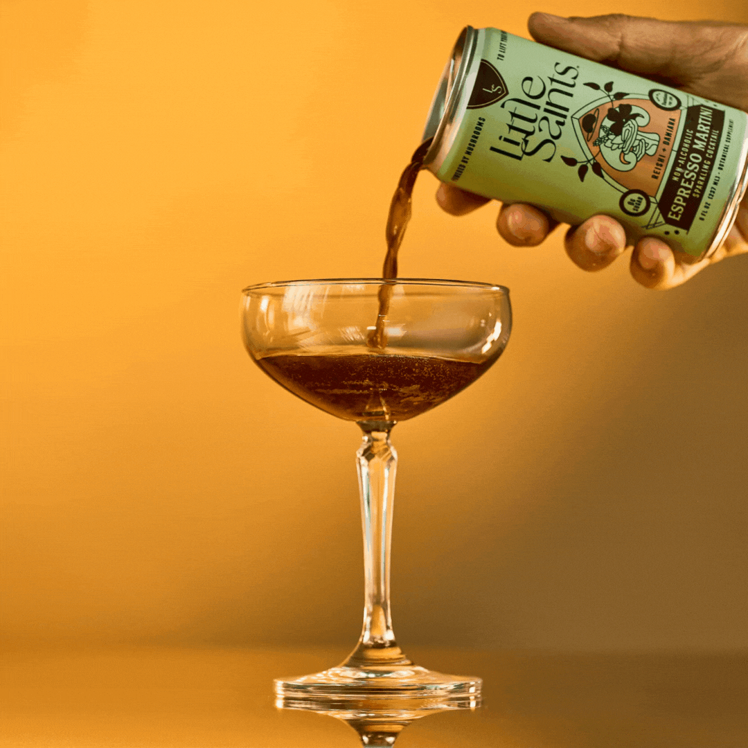 A hand pours Little Saints Espresso Martini Adaptogenic Mocktail from a can into a coupe glass against a warm yellow background.