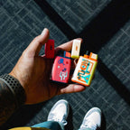 A person holds two colorful rectangular vape devices next to an MK Lighter Company Ignition Pocket Lighter - Nature Series. Their wrist displays a perforated band, while white sneakers stand on a sunlit, carpeted floor.