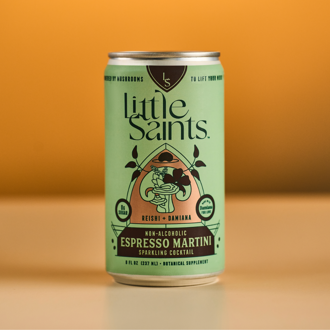 A green can of Little Saints Espresso Martini Adaptogenic Mocktail rests on a light surface against a warm orange backdrop, its label featuring vibrant designs and spotlighting adaptogens like reishi mushroom and Damiana.