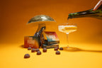 A hand pours sparkling liquid into a coupe glass next to a mushroom disco ball, Fun Guy Flow Fruit Chews boxes, and colorful chews—all set against a vibrant orange backdrop, celebrating these natural mood-enhancing treats.