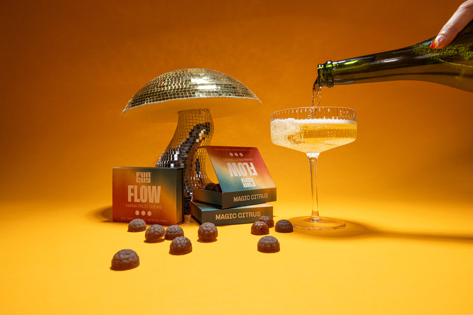A hand pours sparkling liquid into a coupe glass next to a mushroom disco ball, Fun Guy Flow Fruit Chews boxes, and colorful chews—all set against a vibrant orange backdrop, celebrating these natural mood-enhancing treats.